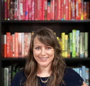 Photo of a white woman with light brown hair in front of a rainbow-sorted bookshelf