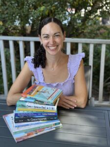 Laurel Symonds sitting at a table with her arms crossed in front of a pile of books she represents.