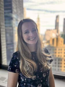 Smiling white woman in an office. Downtown New York is visible through the window behind her.