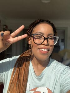 Profile photo of Amanda, a Black woman with ginger braids, black eyeglasses, and blue tshirt, right hand in a peace sign and smiling