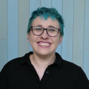 Headshot of Emily. She is a white woman with blue hair and purple glasses and is wearing a black dress.