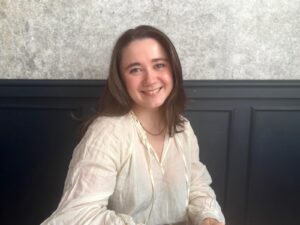 Paper Literary Associate Literary Agent Kiya Evans' headshot - a brunette white woman in a white blouse smiles against a navy and grey backdrop.