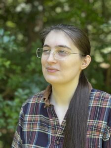 A photo of GiannaMarie, a young white woman with long brown hair and frameless glasses, smiling slightly and wearing a plaid shirt.