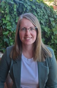 Headshot of a woman against a plant background. She's wearing a green blazer and a white shirt.